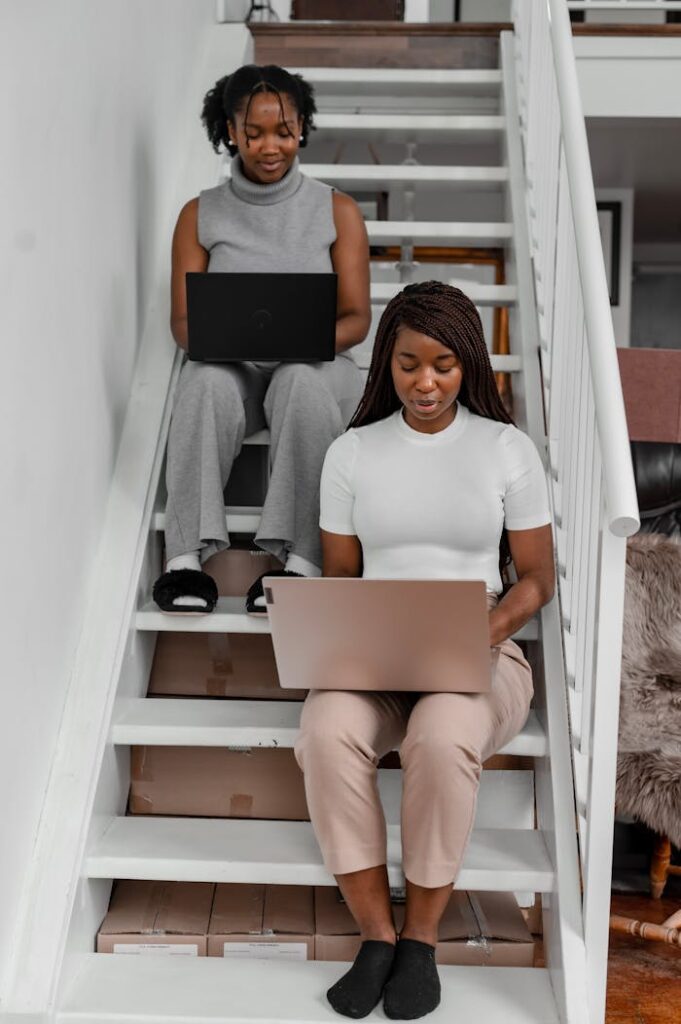 Two women working remotely on laptops from home sitting on stairs indoors.